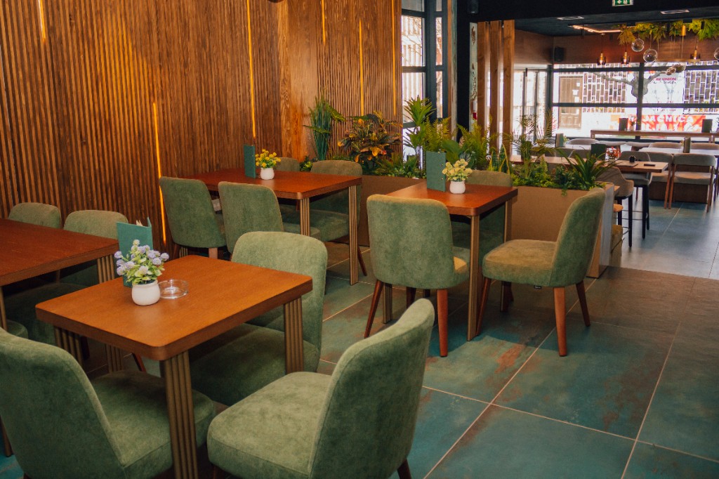 Enterijer sa drvenim stolovima, stolicama u sivo-zelenoj boji, saksijama i toplim osvetljenjem.