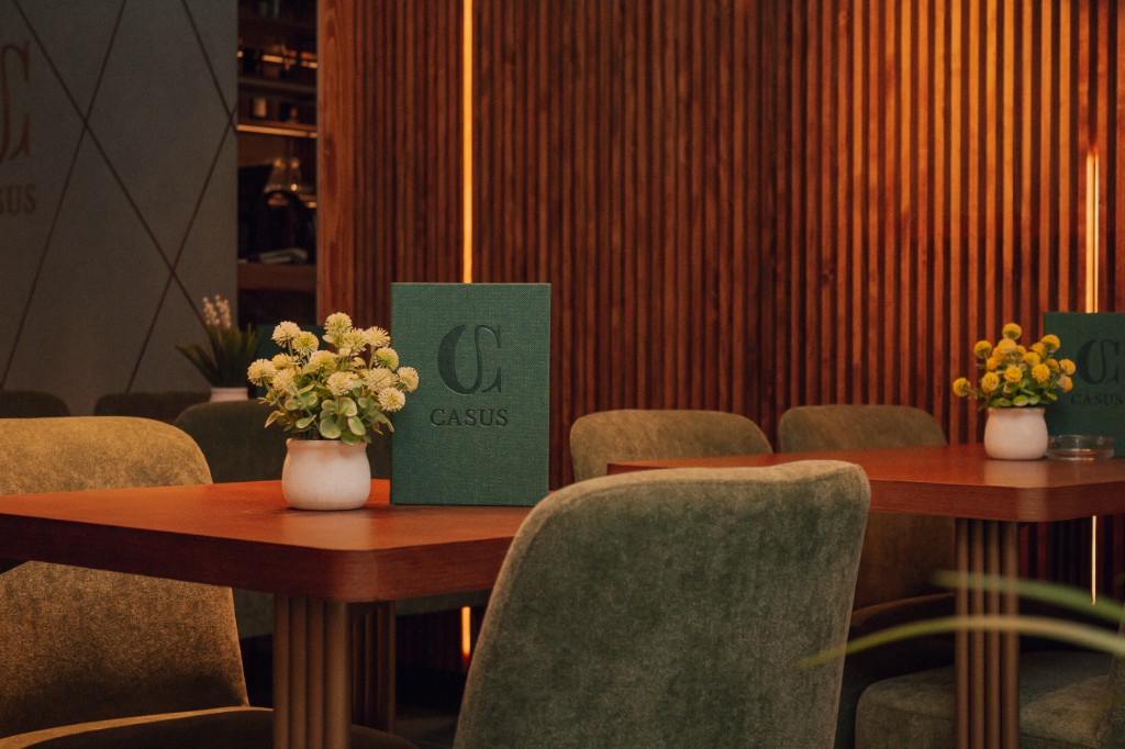 Postavljen sto sa cvećem i menijem Casus, zelene stolice, drveni zid od letvica.