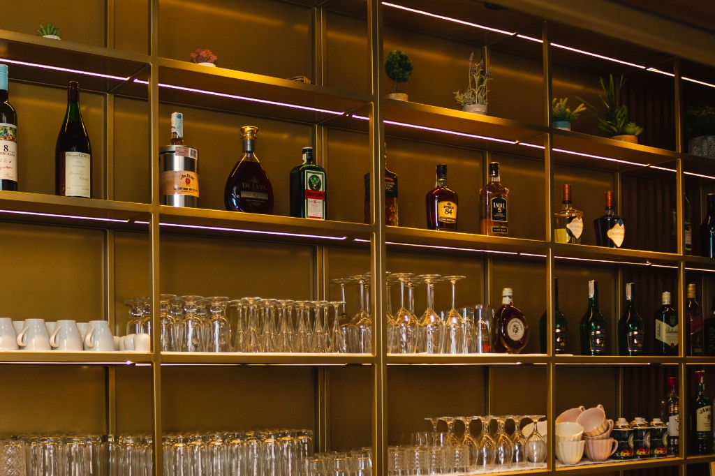 Šank sa zlatnim policama, bocama vina i žestokih pića, posuđem.