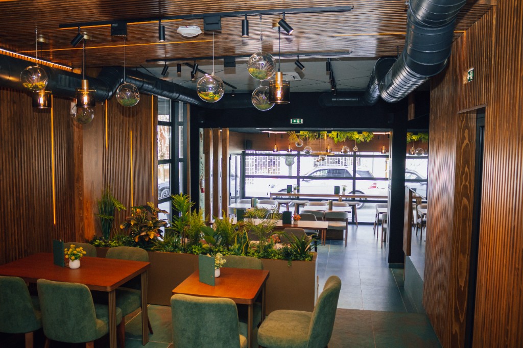 Prostor za jelo sa drvenim stolovima, zelenim stolicama, saksijama i staklenim lustrama.